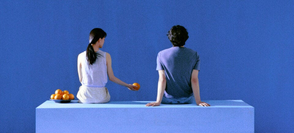 Two people sitting on a blue surface against a blue background, with a bowl of oranges beside them.