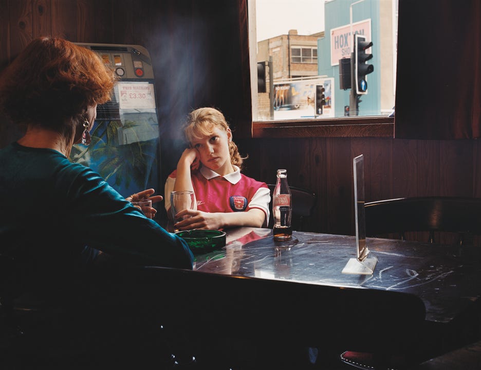 A girl in a pink vest sits at a table in a dimly lit room, looking pensive. Another person smokes beside her.