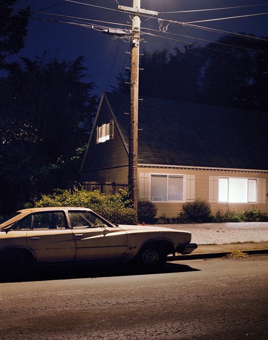 Old car parked on street at night, illuminated by streetlight. House with lit windows in the background.