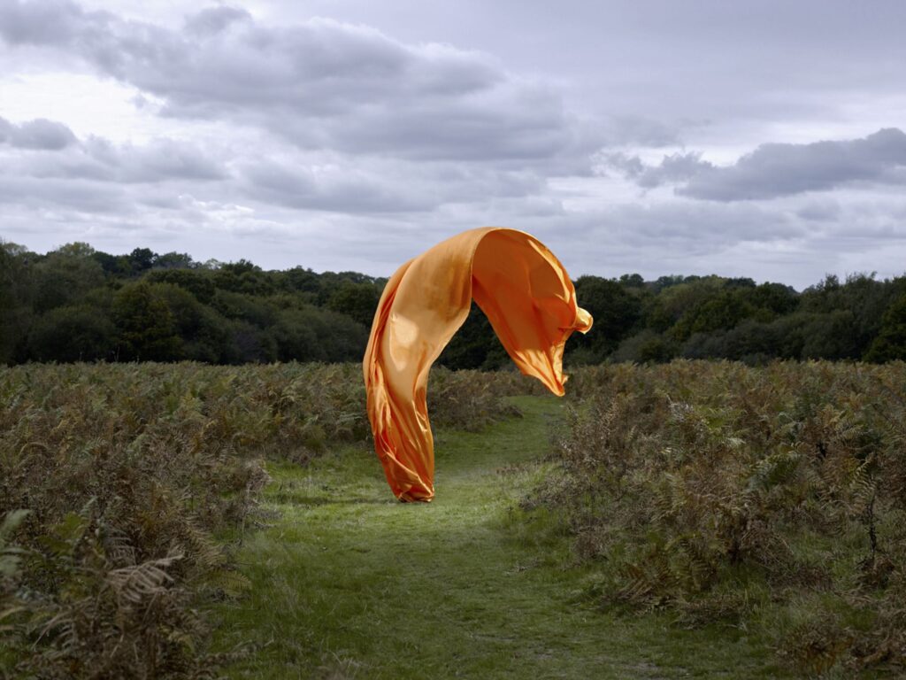 Orange fabric floats mid-air over a green field with cloudy sky in the background.
