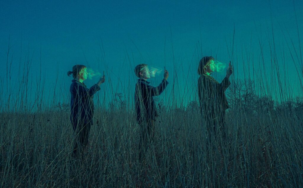Three people in a field face smartphones, glowing screens illuminate their faces, creating a surreal technology contrast.