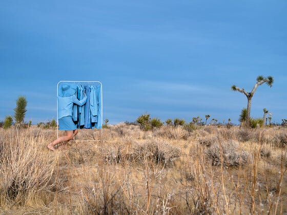 Person pushing a rack of blue clothes through a desert landscape with sparse vegetation under a clear sky.