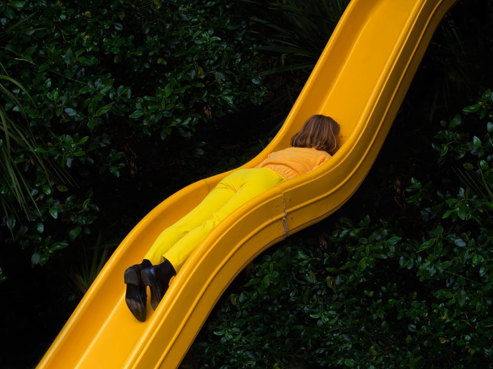 Person in yellow outfit sliding headfirst down a bright yellow slide surrounded by green foliage.