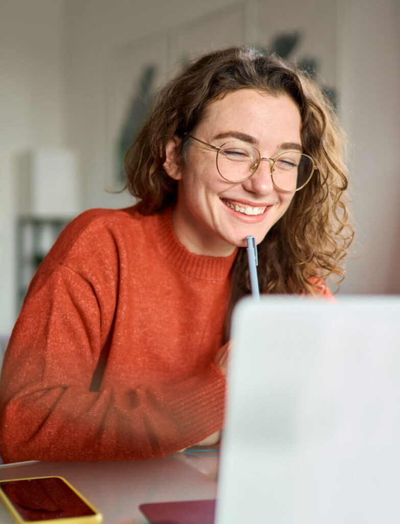 Person with curly hair and glasses in a red sweater smiling at a laptop screen, holding a pen to their chin.