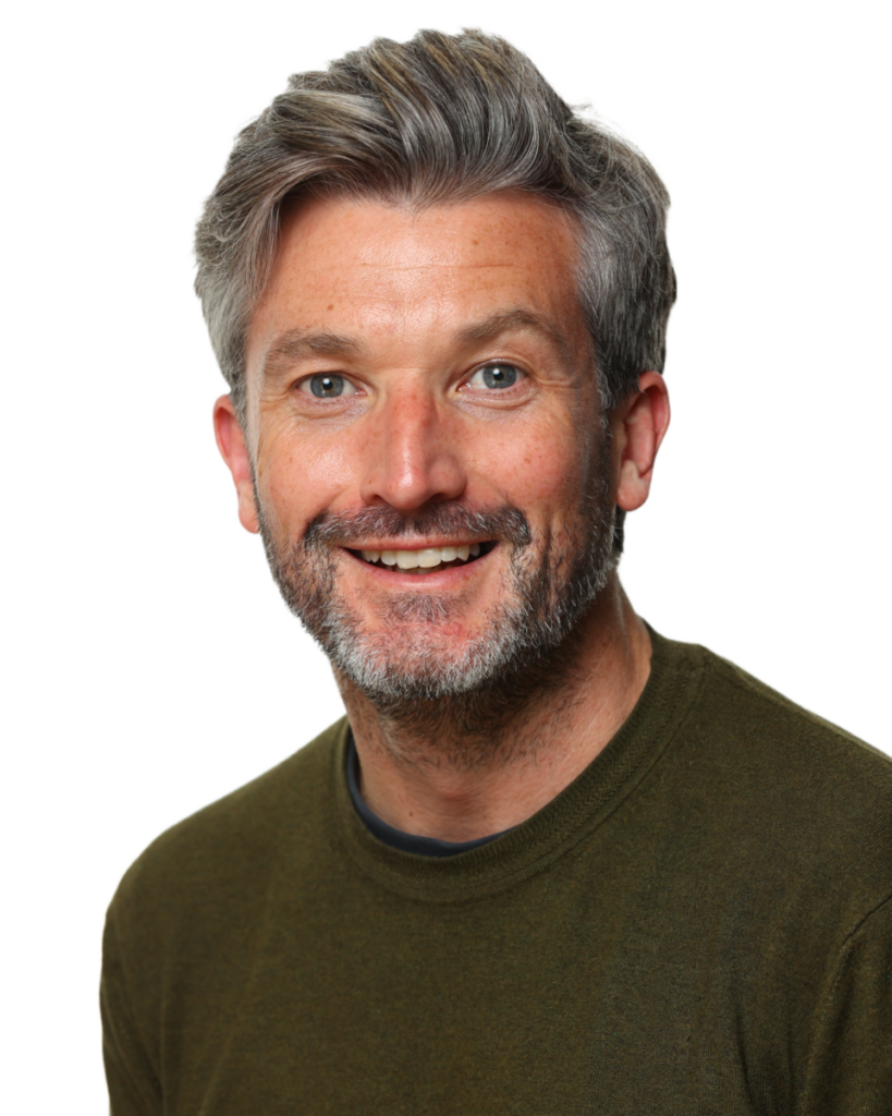 Smiling man with grey hair and beard wearing a dark green sweater against a plain white background.