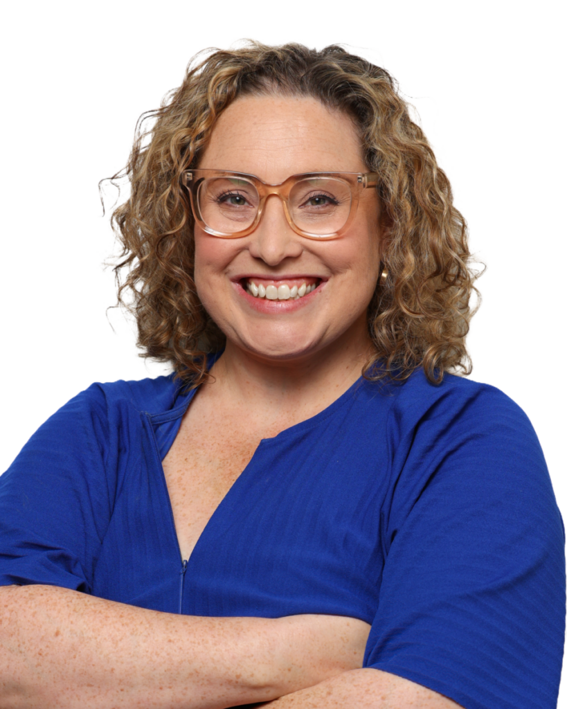 A smiling person with curly hair and glasses, wearing a blue top, crosses their arms confidently.