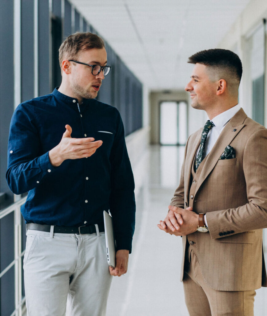 Two men in a hallway having a conversation; one wears a blue shirt, the other a beige suit.