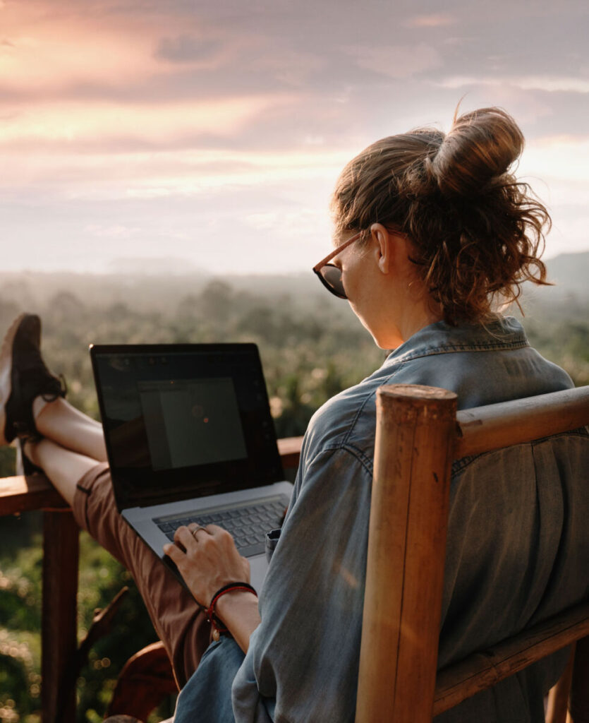 Person with sunglasses using a laptop on a balcony, overlooking a lush landscape at sunset.
