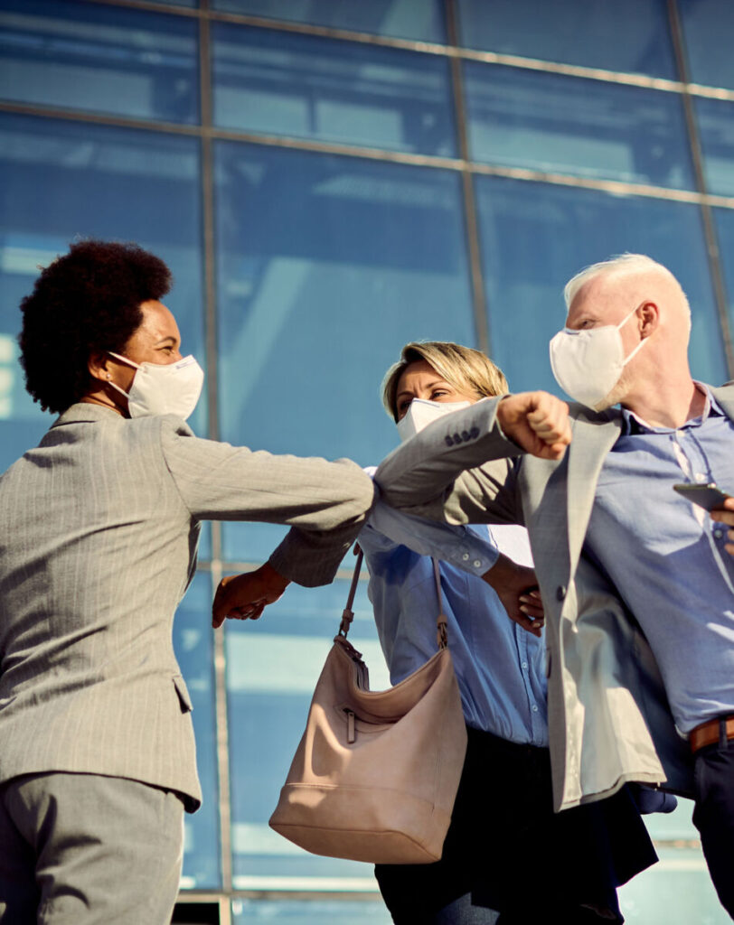 People wearing masks, greeting with elbow bumps outside a modern glass building.