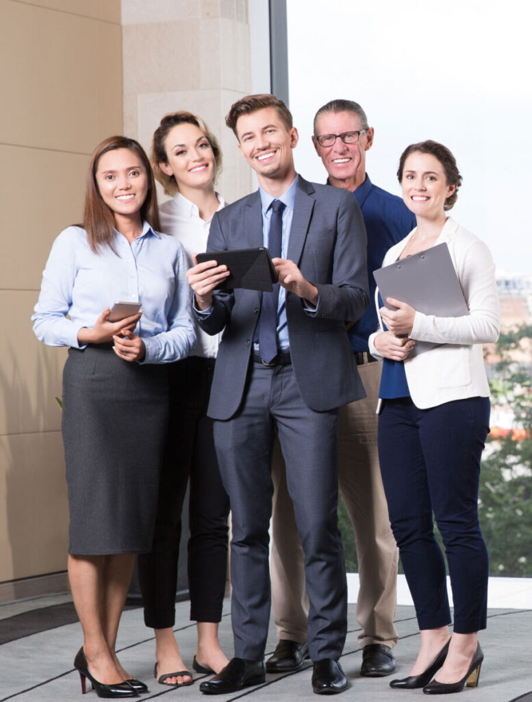 A diverse group of business professionals smiling and holding devices, standing together in an office setting.