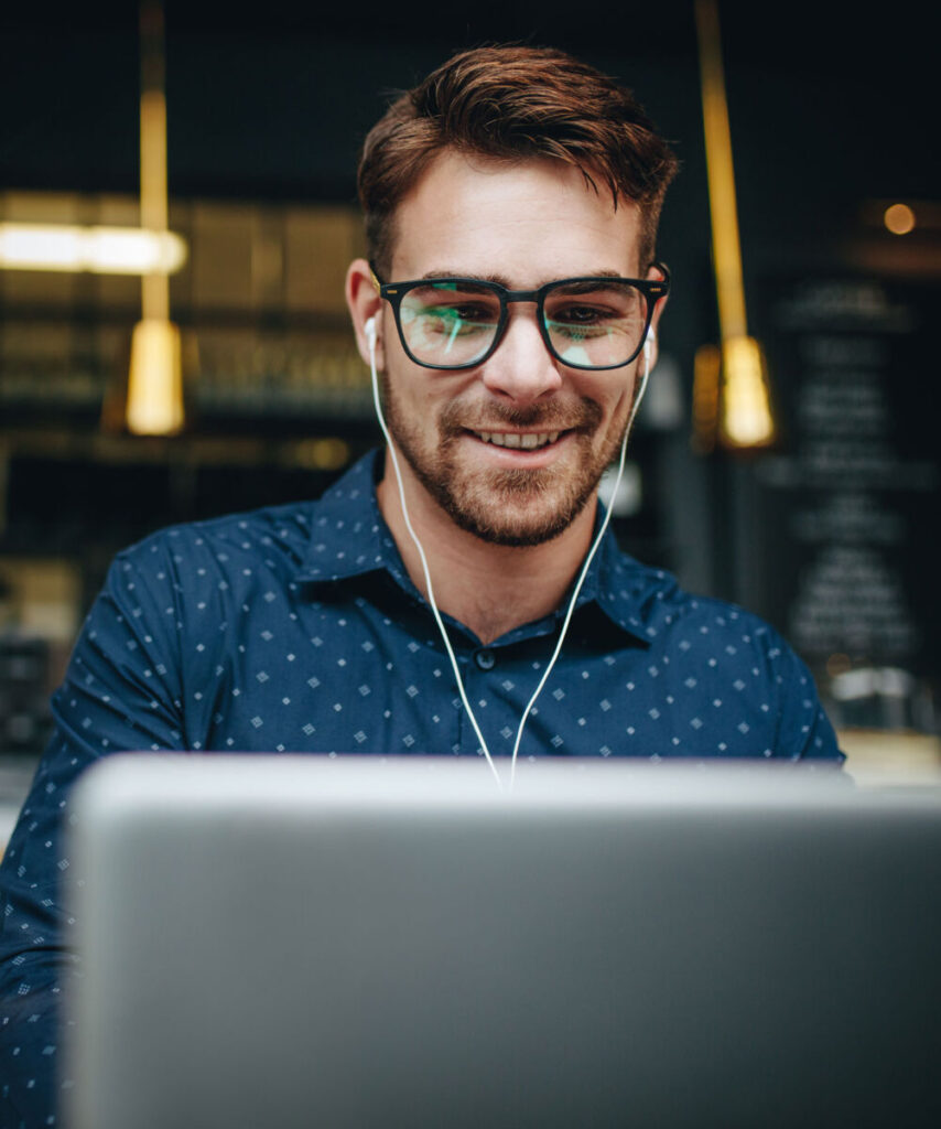 Young man with glasses and earphones smiling while using a laptop in a cozy café setting.