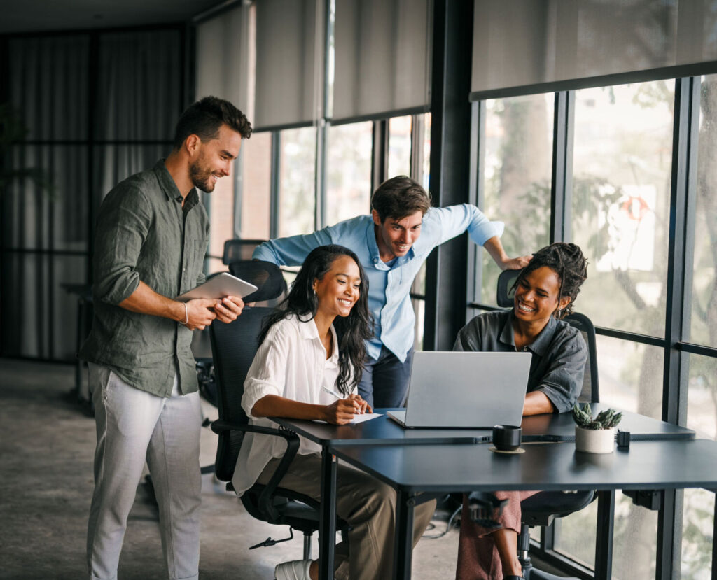 A diverse group of four colleagues collaborate joyfully around a laptop in a modern office with large windows.
